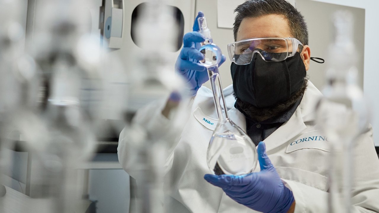  A Corning employee wearing a lab coat, gloves, and safety glasses holds a glass flask in front of a frosted glass background.