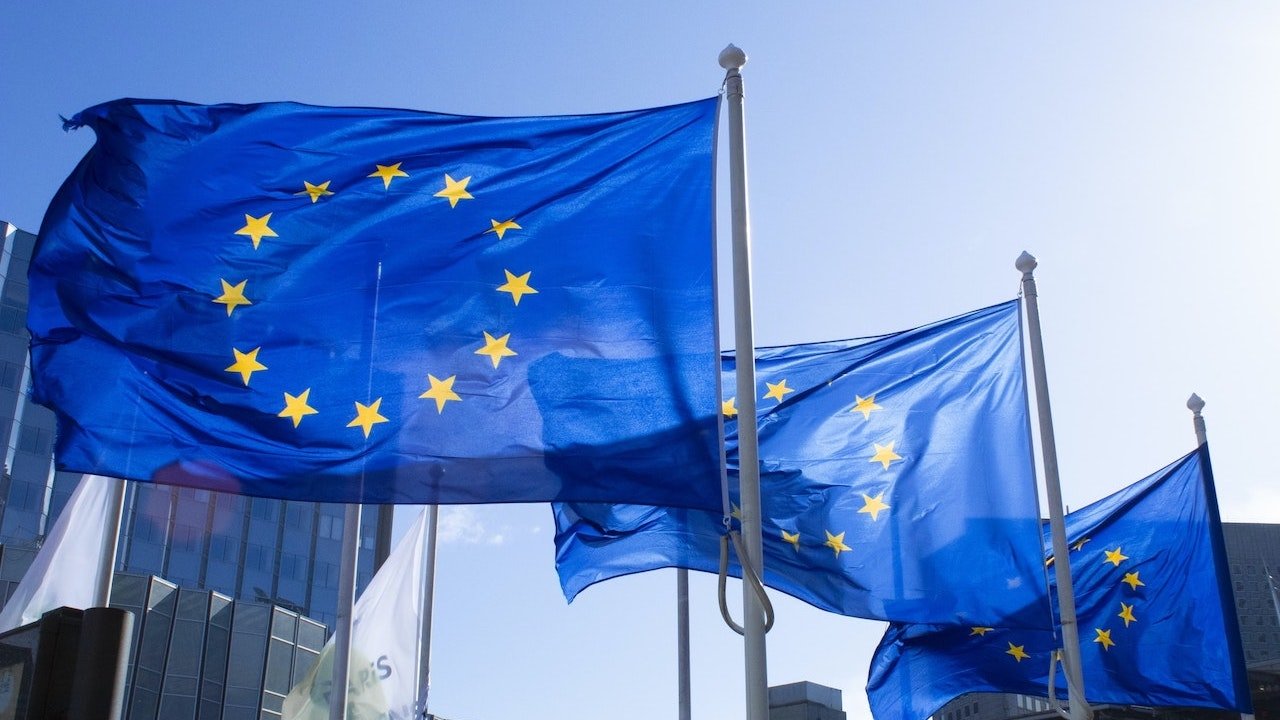 Three European Union flags fluttering in front of two buildings and under blue sky