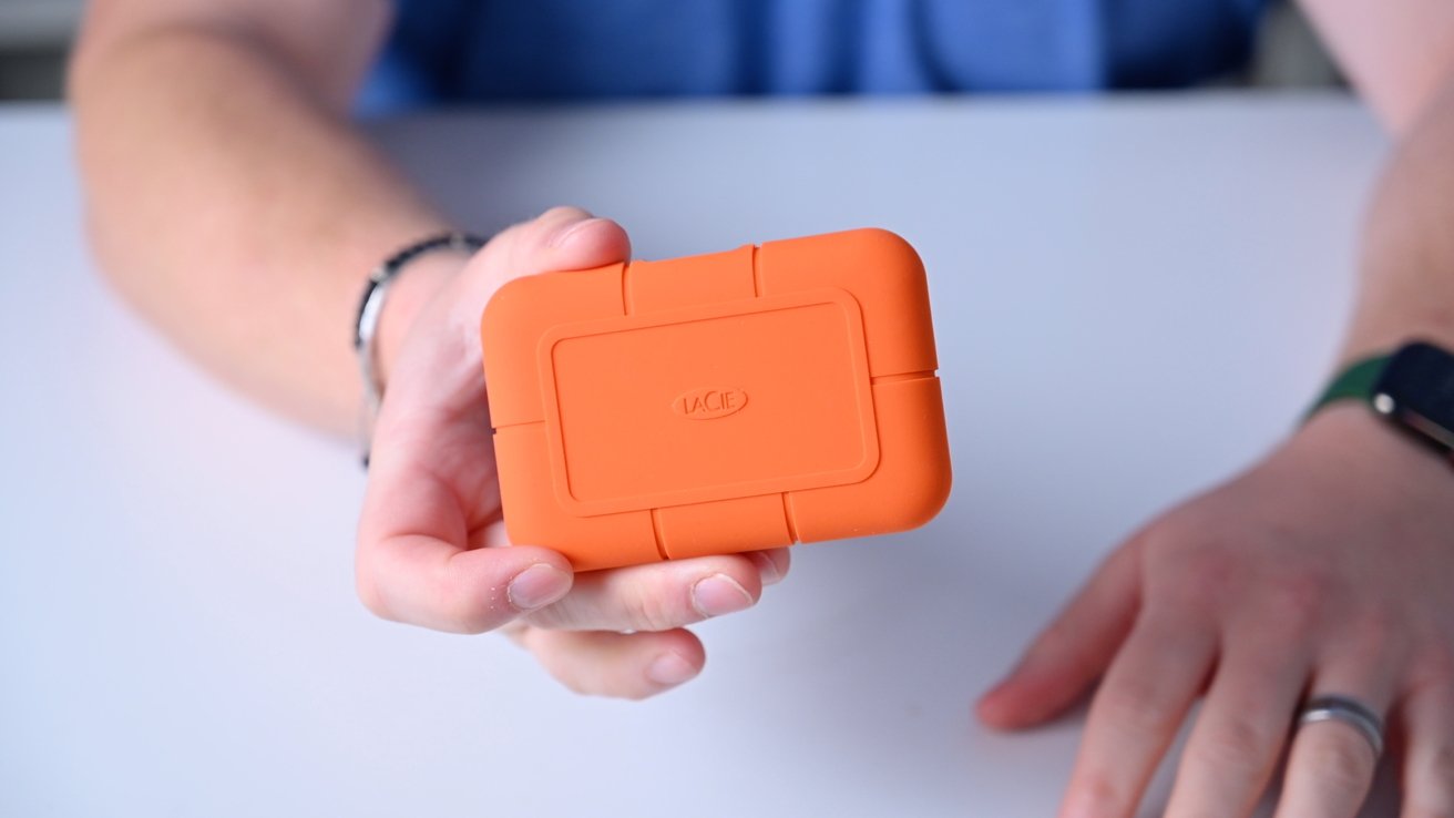 A person wearing a smart watch holds an orange, rugged external hard drive close to the camera.