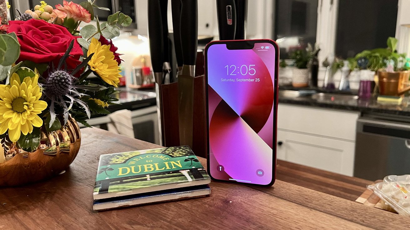 iPhone 13 refurbished models offer cost savings compared to buying the iPhone 14 | Phone in red on kitchen counter next to book