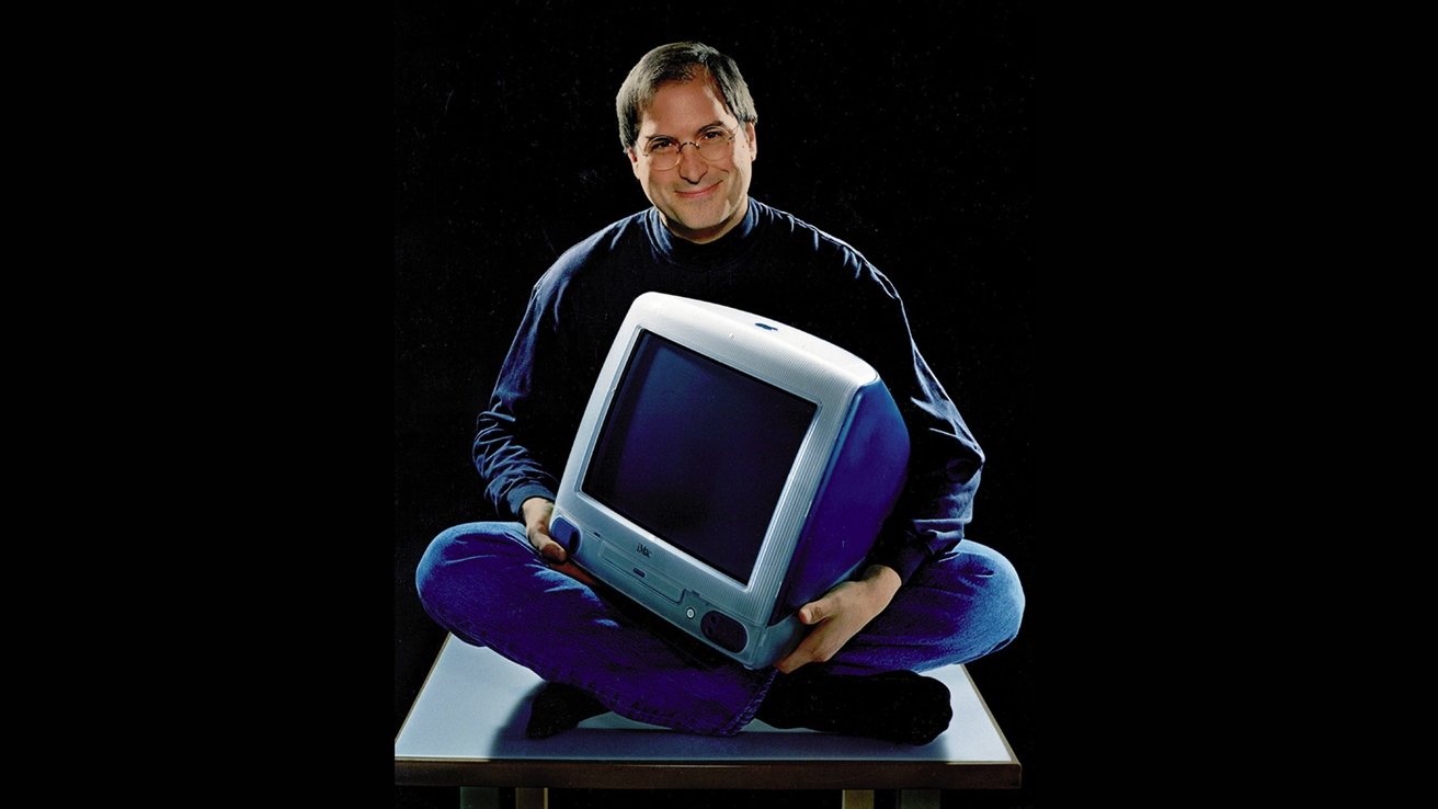 A smiling Steve Jobs holding the original iMac while sitting cross-legged on a platform against a dark background.