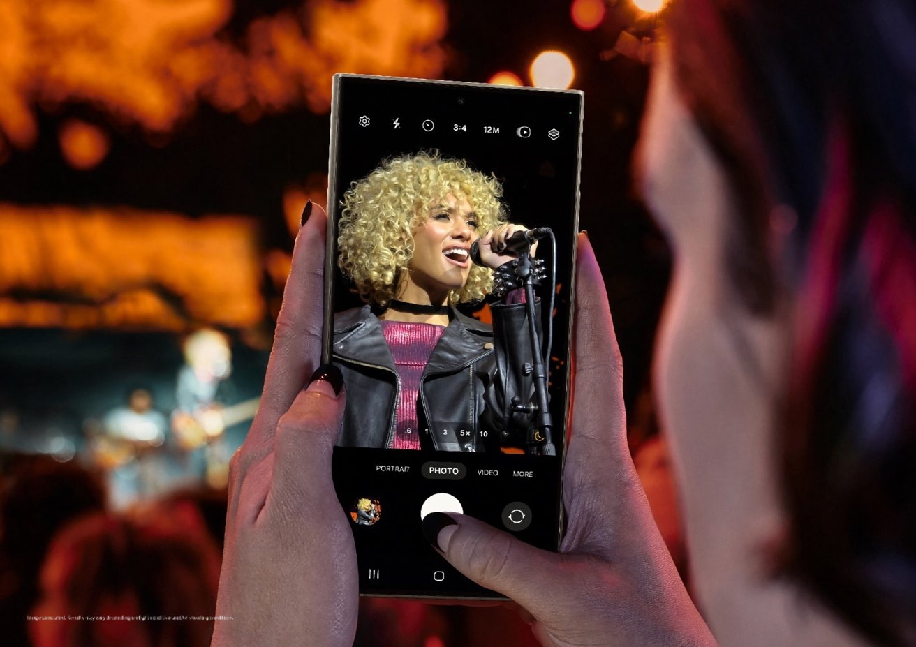 Hands holding a smartphone capturing a singer performing onstage, seen through the phone's camera screen, with a blurred audience in the background.