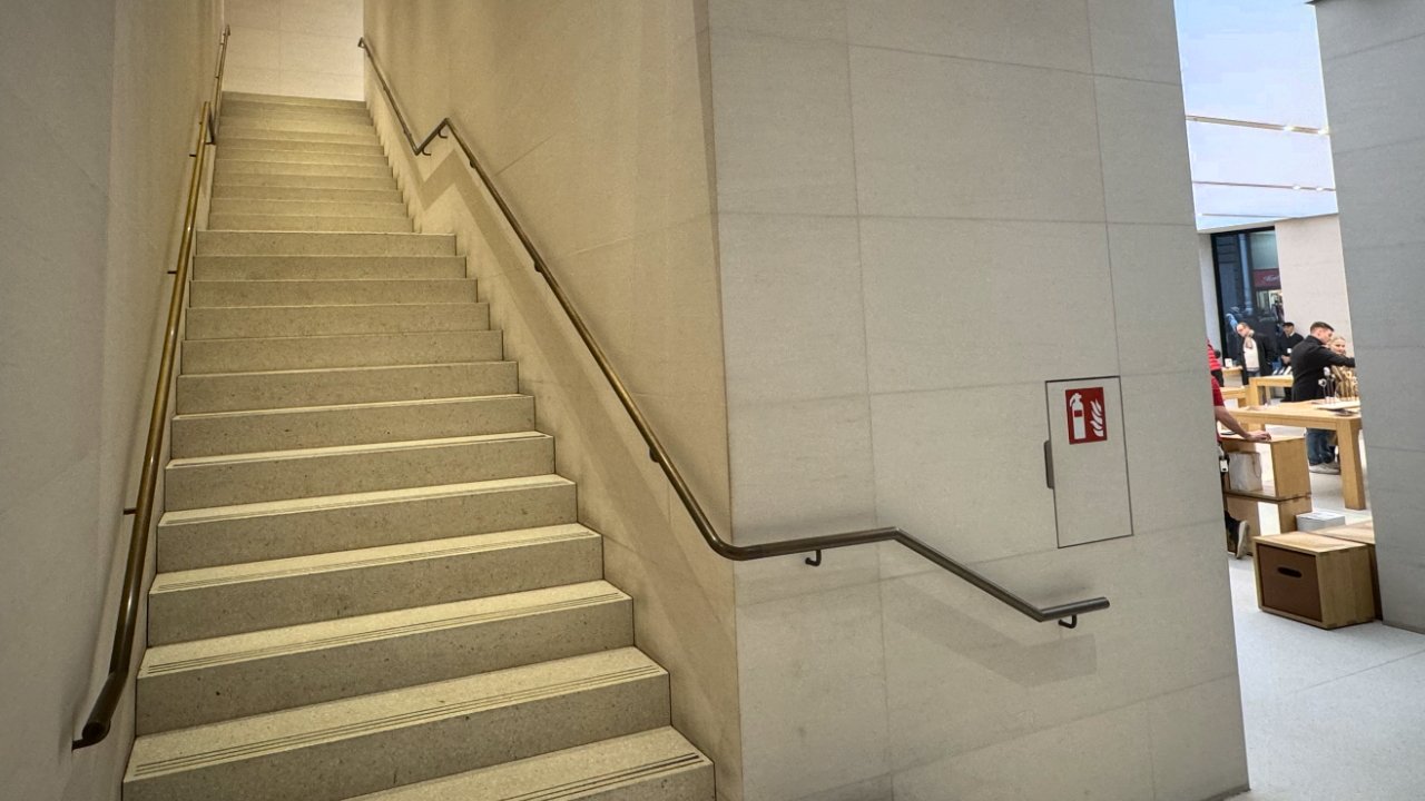 A staircase in Apple Karntner Strabe, with beige steps leading up to a landing with a metal handrail on the right and a fire extinguisher sign on the adjacent wall.