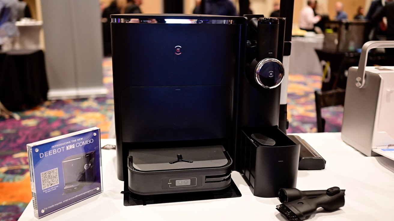 A sleek, black robotic vacuum cleaner with a detachable hand vacuum and accessories on display, alongside promotional material on a table at a technology event.