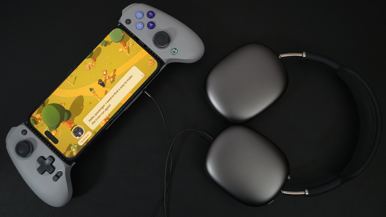 iPhone in a GameSir controller shows a scene from 'Tamagatchi,' a colorful game with a yellow character walking through woods. A pair of gray AirPods Max lie next to the controller.