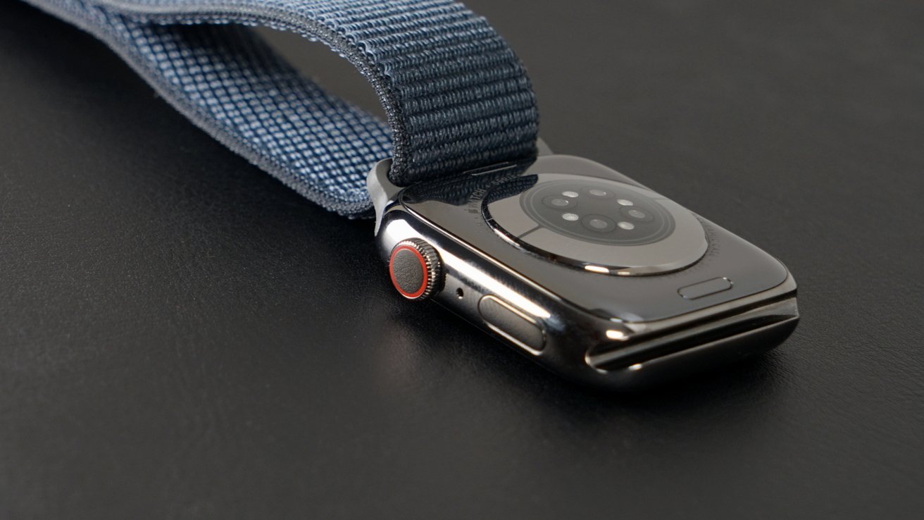 An Apple Watch with a navy blue fabric strap lies on a black surface; its digital crown has a red accent. The underside of the watch, with sensor array and charging coils, is visible.