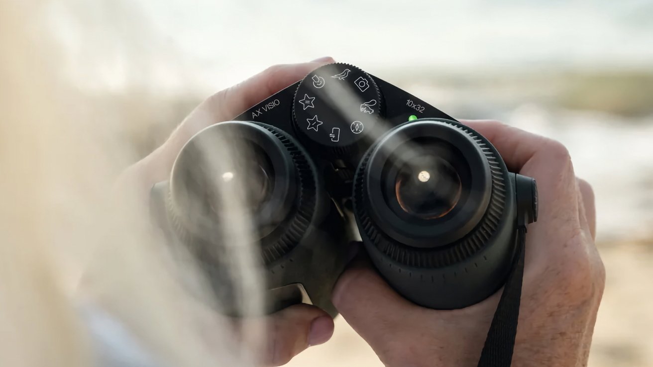 A pair of black binoculars held by a human hand against a blurry natural backdrop, focusing on the dial with various mode symbols on top.