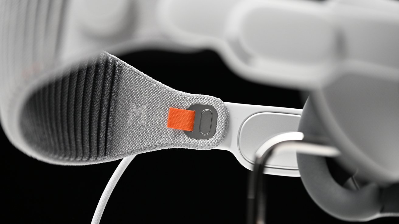 A close-up view of the Apple Vision Pro headset strap with fine texture, a gray fabric, and an orange adjustment tab, against a blurry white background.