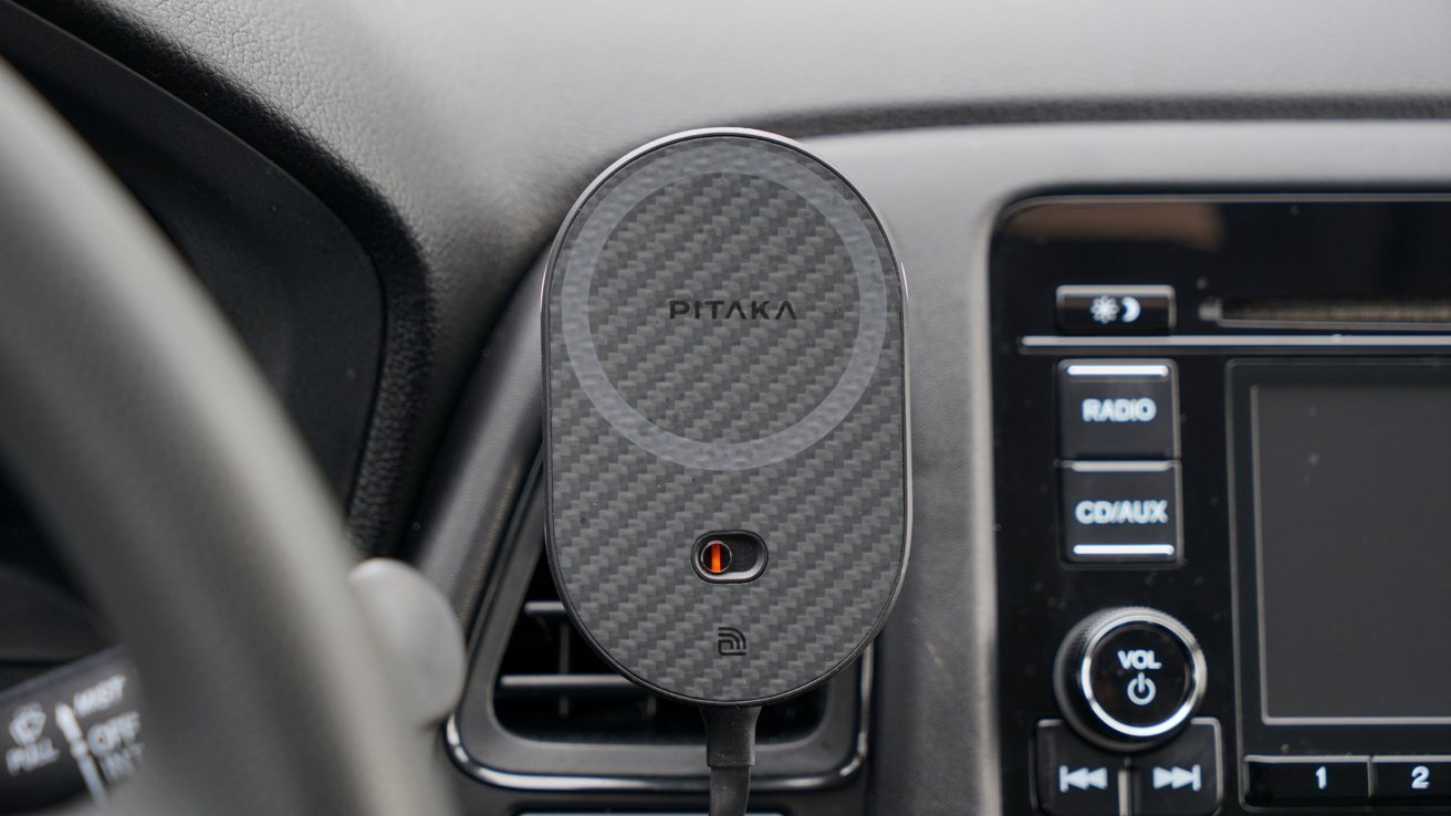 A carbon-fiber-patterned PITAKA magnetic car mount attached to a vehicle's dashboard next to the infotainment system display.