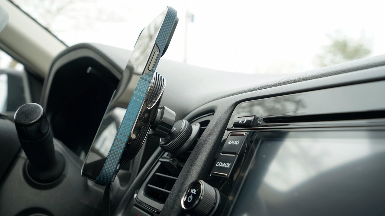 A smartphone mounted on a car's air vent holder beside the dashboard, with various car control buttons like radio and hazard lights visible.