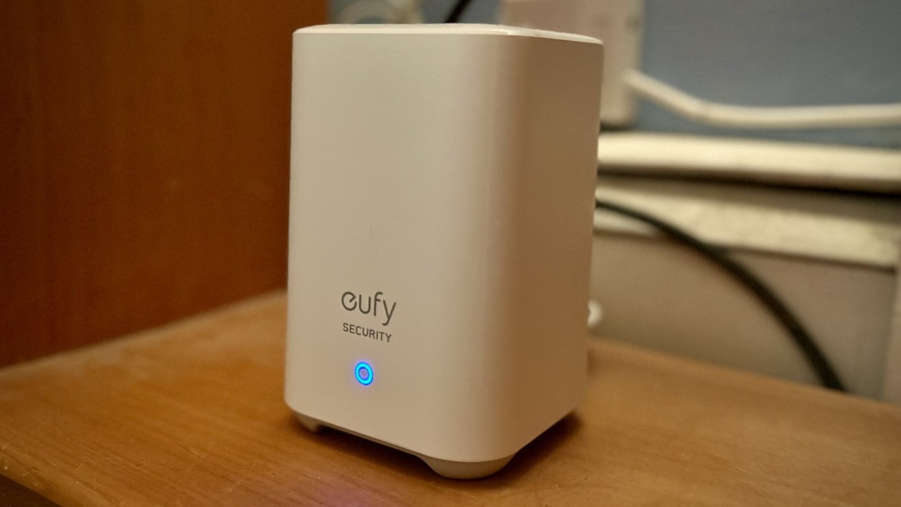 A white rectangular electronic device labeled 'eufy SECURITY' with a glowing blue circular light near its base, set on a wooden surface against a blurred indoor background.