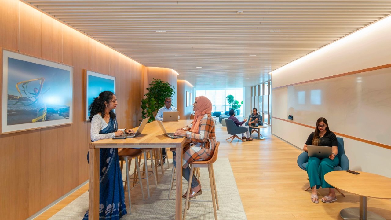 Apple's modern office space in Bengaluru with wooden walls and people working at laptops, some seated at desks and others in casual seating areas. Large framed photos decorate the wall.