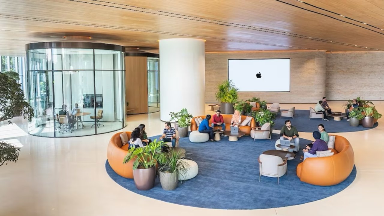 Apple's modern office lobby in Bengaluru, India with people seated on sofas and chairs around circular planters, under warm lighting. In the background, a large Apple logo on a screen, and a glass-walled meeting room to the left.
