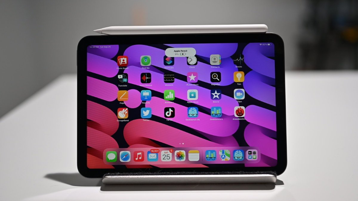 An iPad mini stands on a white surface with a vibrant, colorful display showing app icons. A stylus rests on top, indicating touchscreen functionality.