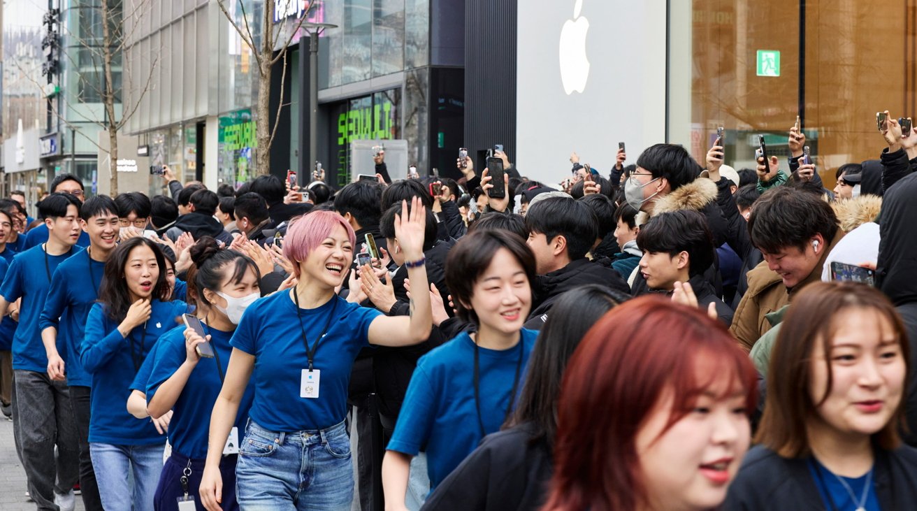 A crowd of enthusiastic people, many holding up smartphones, gathers outside an Apple store, with employees in blue shirts smiling and high-fiving guests.