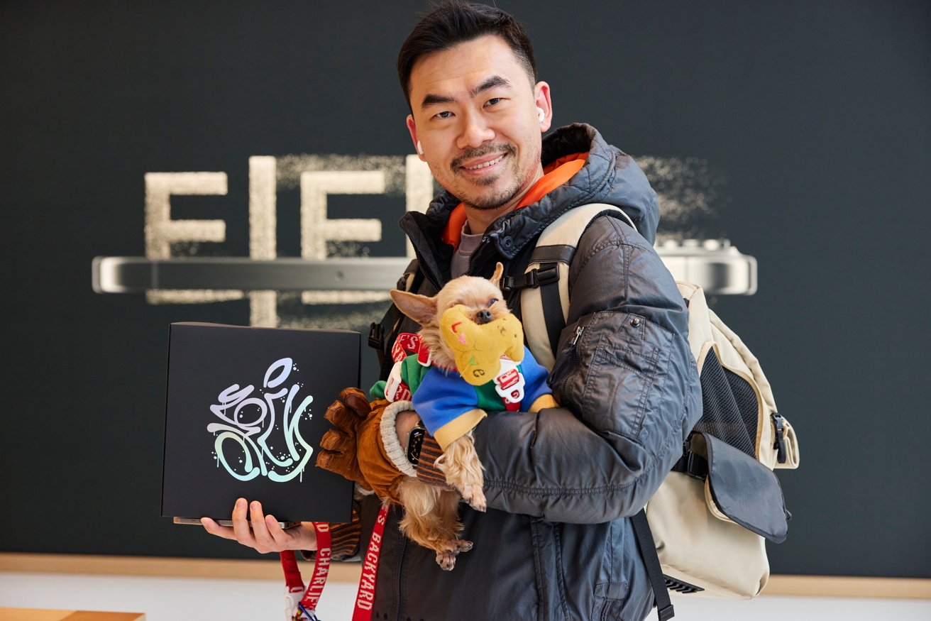 A smiling man holds a decorated box at an Apple Store in South Korea. He is wearing a jacket and backpack, with a lanyard around his neck.