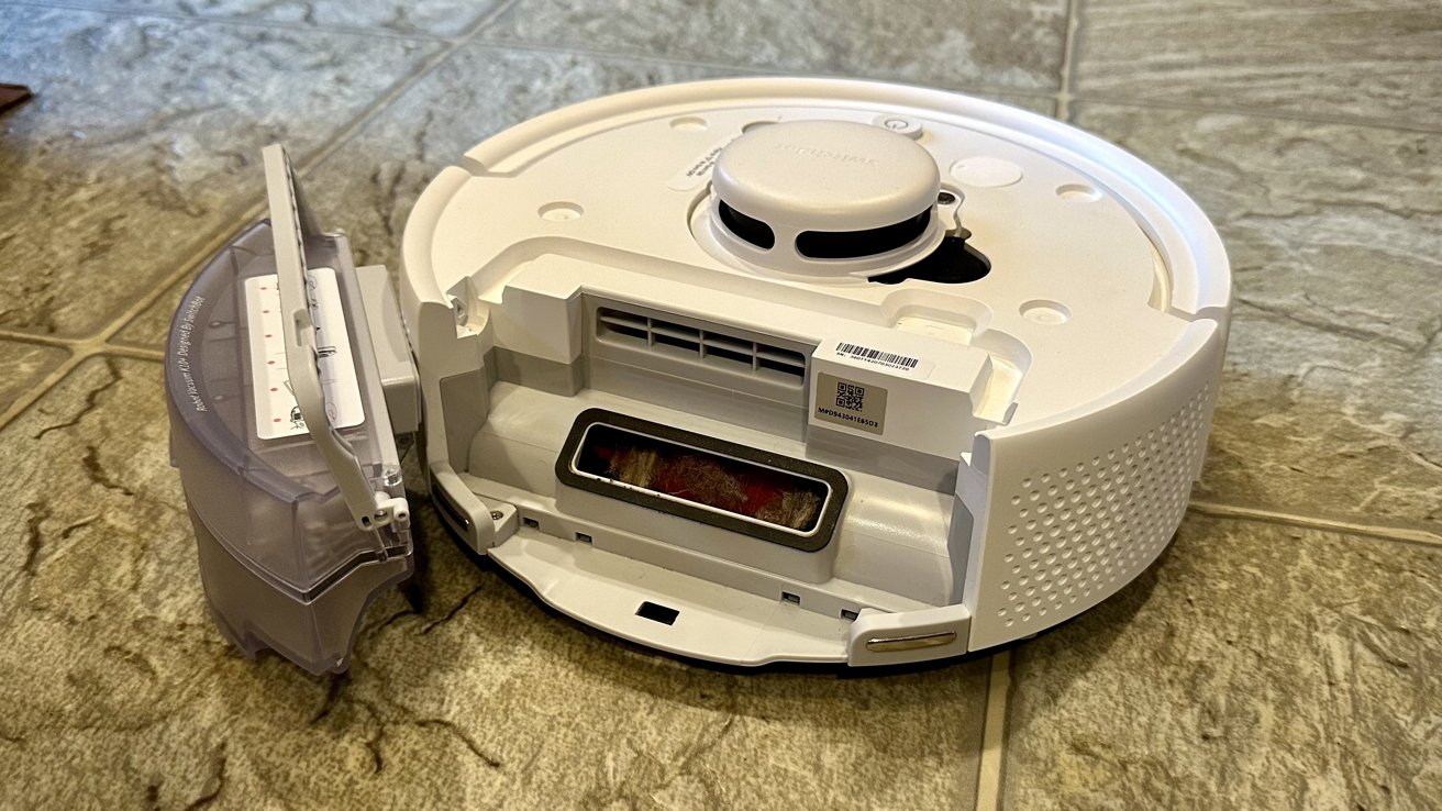 An opened robotic vacuum cleaner on a tiled floor with its dustbin and brush exposed.