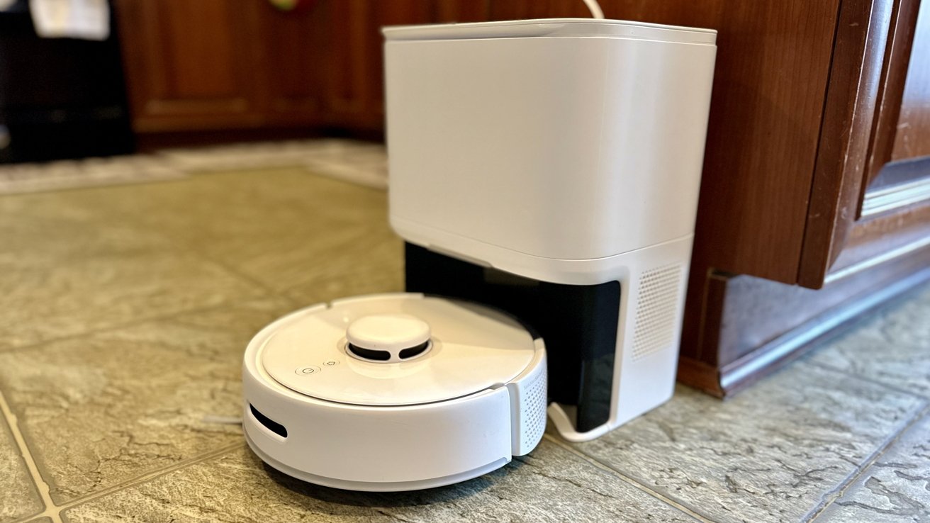 A white robotic vacuum docked at its charging station on a tiled floor near a wooden cabinet and a glass door.