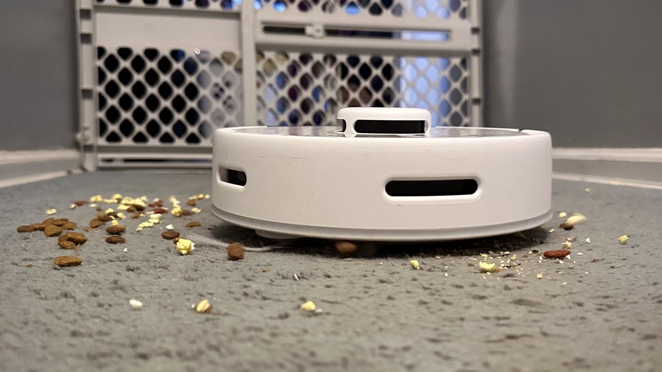 A robotic vacuum cleaner surrounded by scattered pet food and crumbs on a carpeted floor.
