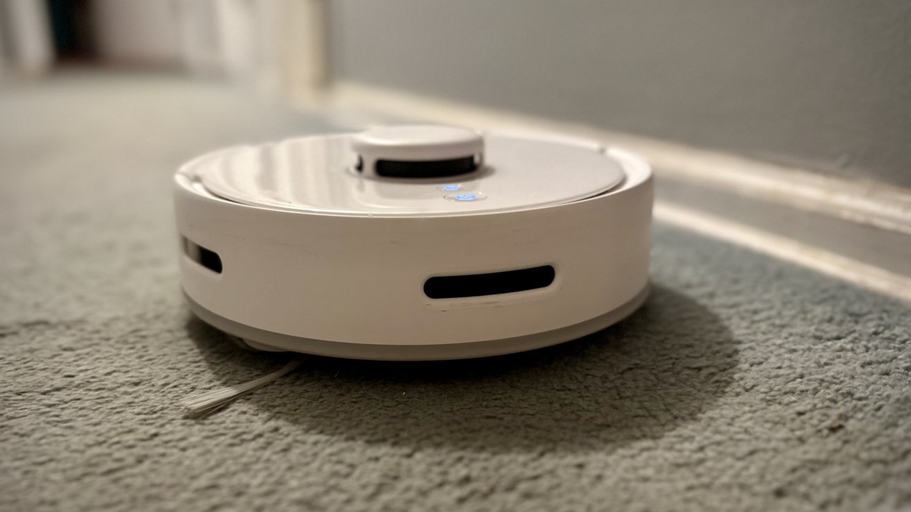 A white robotic vacuum cleaner on a grey carpeted floor near a wall, with illuminated buttons visible on top.