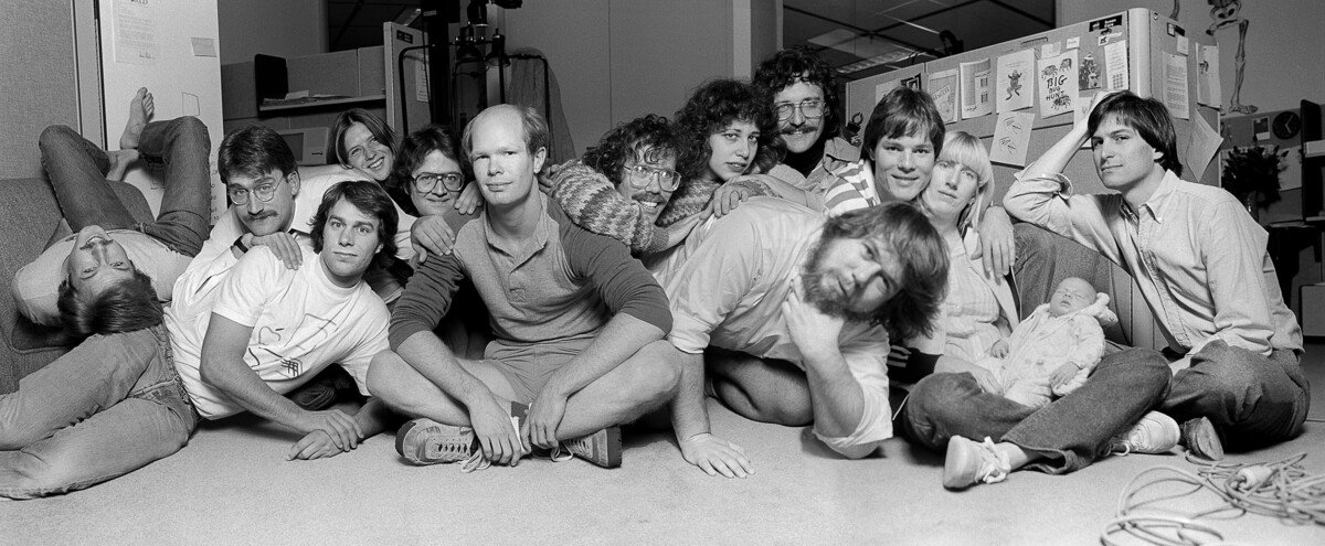 Black and white photo of a group of casually dressed people, including Steve Jobs, playfully posing in an office setting, some seated on the floor, others leaning on cubicles, with one cradling a doll.