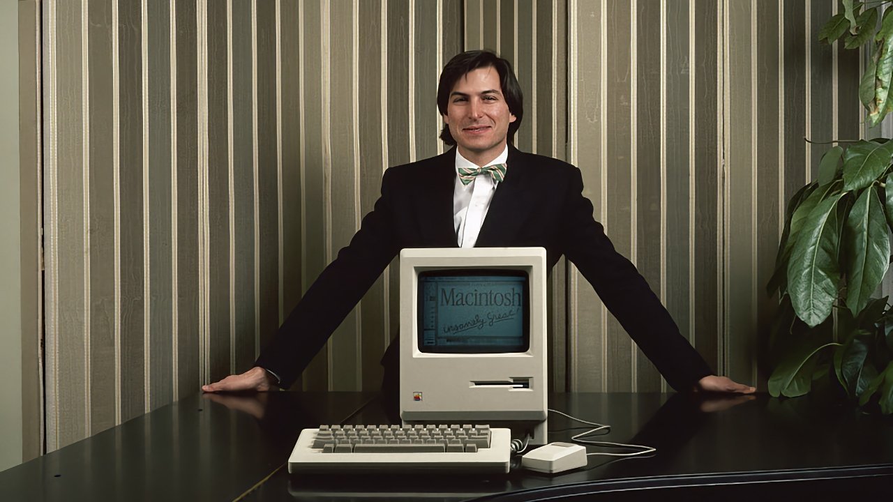 Steve Jobs smiles behind an original Macintosh computer on a desk, with striped wallpaper and plant leaves to the side.