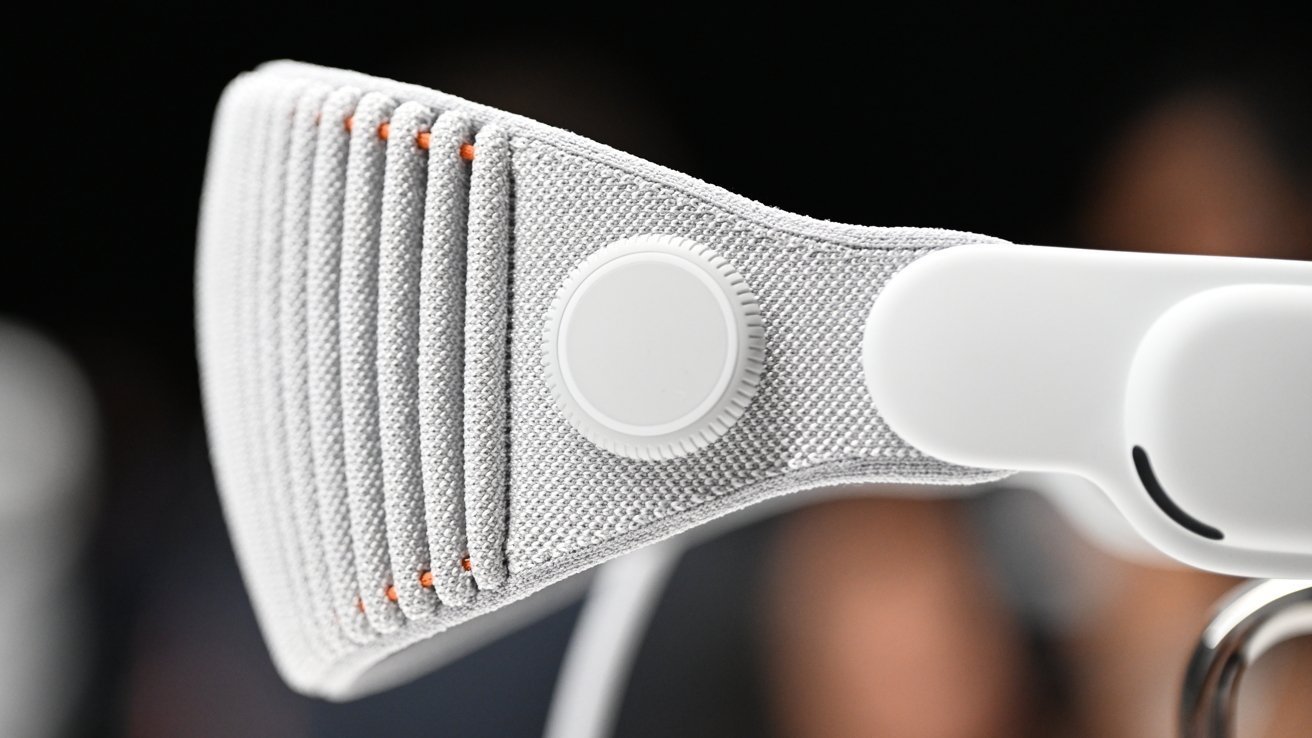 Close-up of a gray fabric headset strap with a circular white knob, accentuated by small orange stitches, focused against a blurred background.