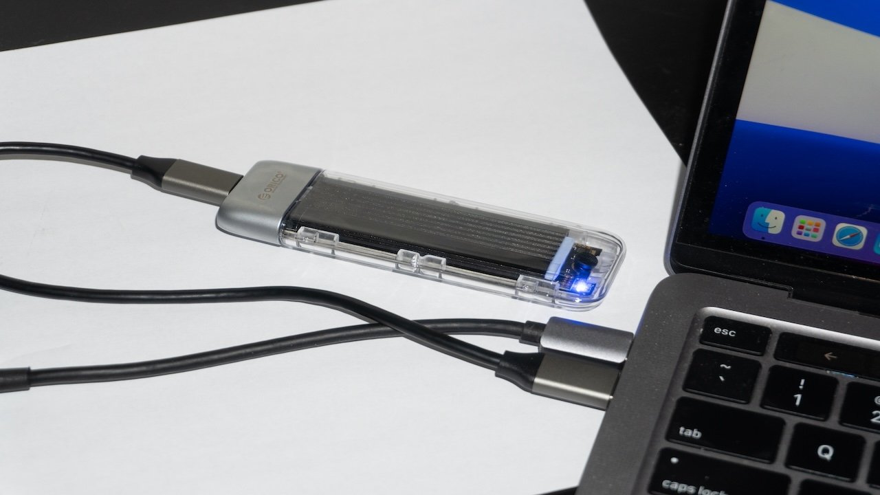 A clear USB SSD connected to a laptop via a cable, with a blue LED light indicating activity, resting on a white surface next to the black keyboard of the laptop displaying colorful icons on its screen.