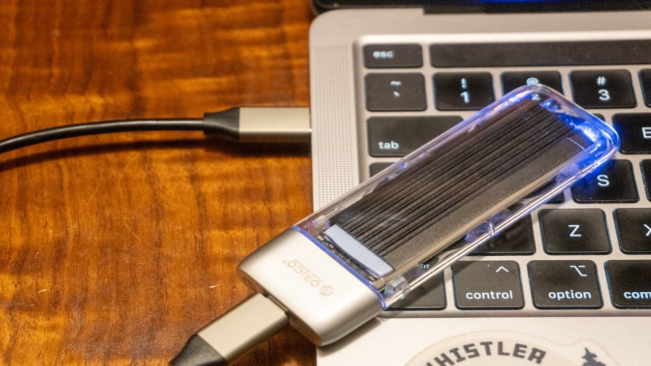 A USB-C dongle connected to a laptop with a glowing external hard drive on a wooden surface.
