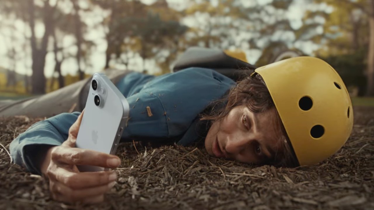 A person wearing a yellow helmet lies on the ground holding out an iPhone 15, with a pained expression on their face. There are trees and yellow leaves in the background, suggesting an outdoor setting.