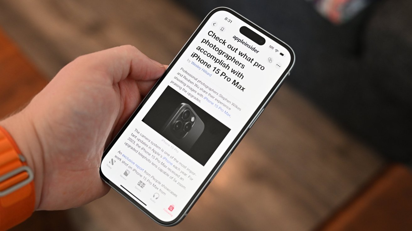 A hand holding an iPhone 15 displaying an article about iPhone 15 Pro Max camera capabilities with a photo of the phone.