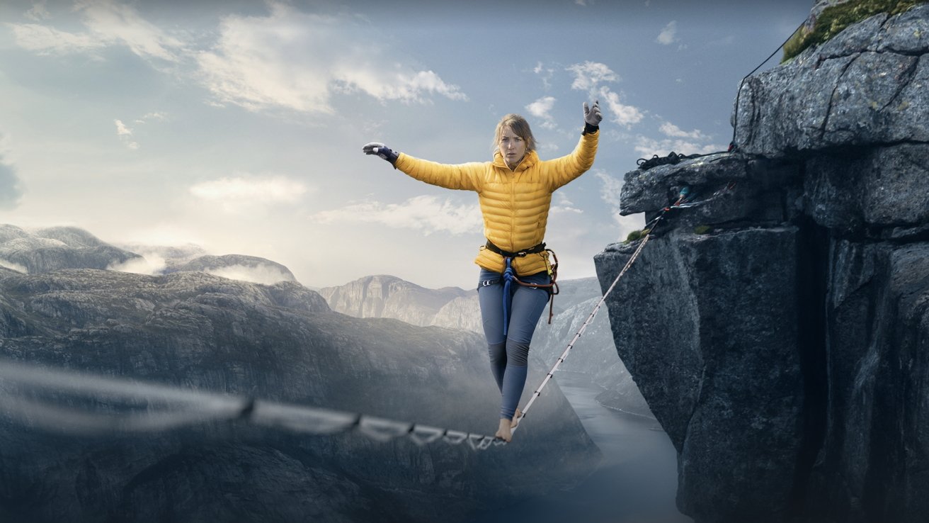 Person balances on a slackline high above a scenic canyon with misty mountains in the background, wearing safety gear and focused expression.