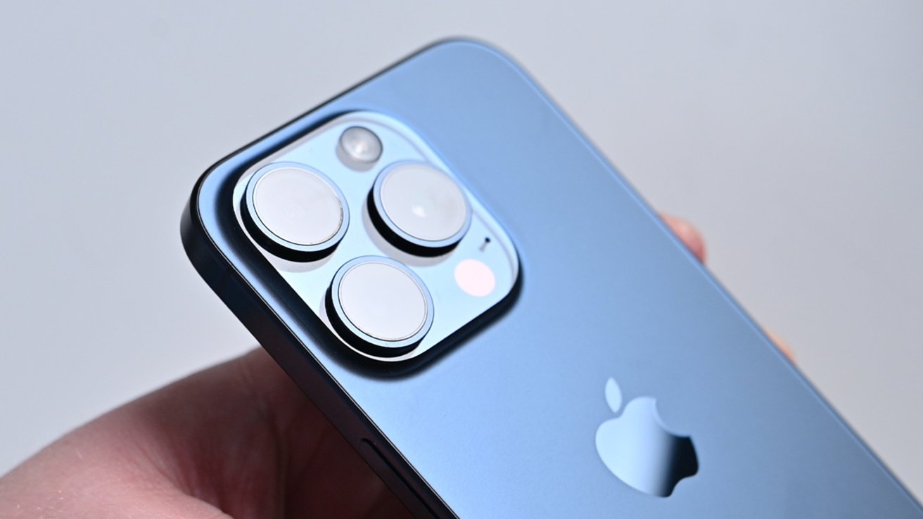 A blue iPhone 15 Pro held in a hand with the three cameras reflecting a nearby light.