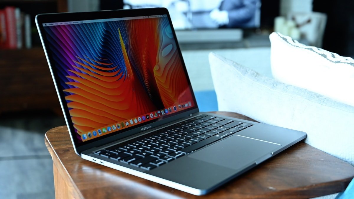 MacBook Pro with colorful wallpaper on screen, on a wooden table, with a person in the blurred background.