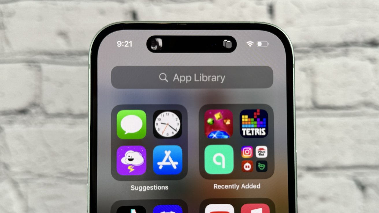 An iPhone shows its App Library with several colorful icons. The device is standing with a blurry white brick background.
