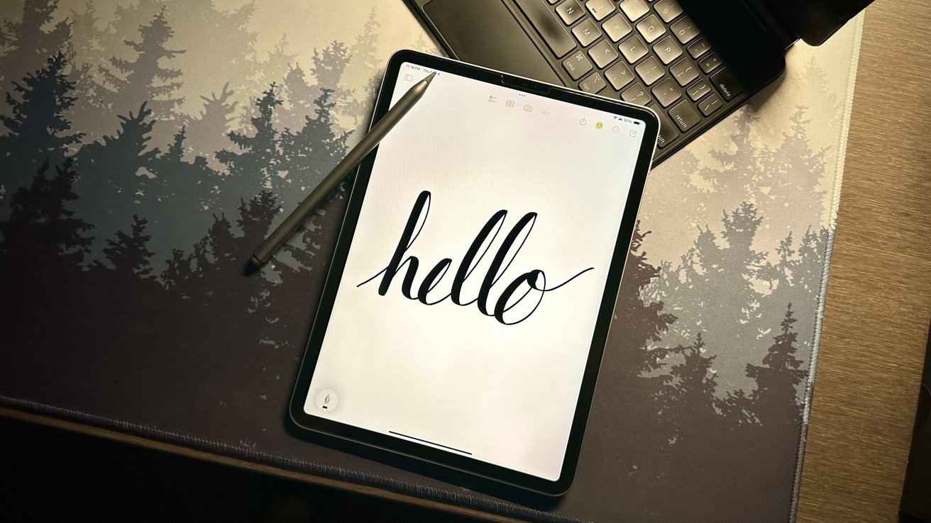 An iPad with stylus displaying handwritten 'hello' on screen, resting on a desk next to a keyboard with tree patterns.
