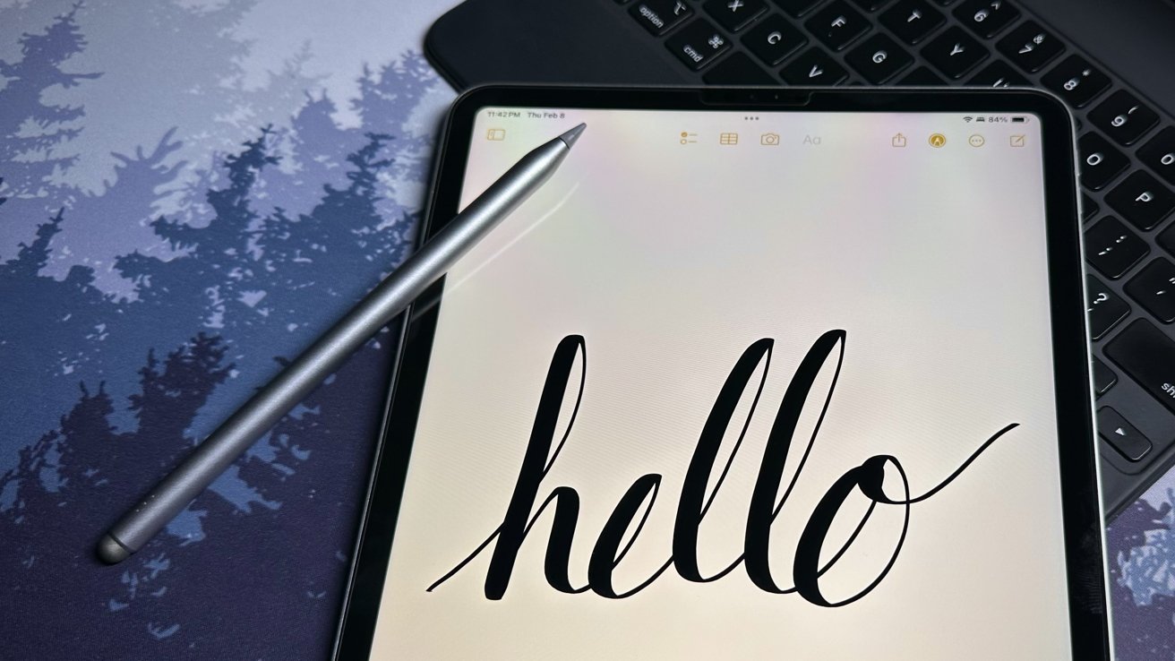 A stylus pen rests on an iPad displaying the handwritten word 'hello' with a keyboard in the background.