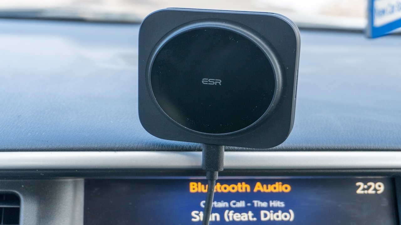 Black square wireless charger mounted on car dashboard, cable connected, with display screen indicating Bluetooth Audio in background.