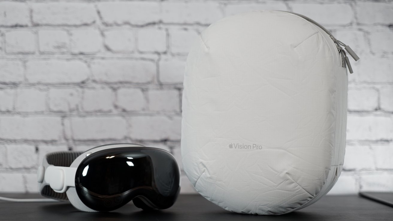 Apple Vision Pro sits next to the large white oval Travel Case on a table. The background is blurred white brick.