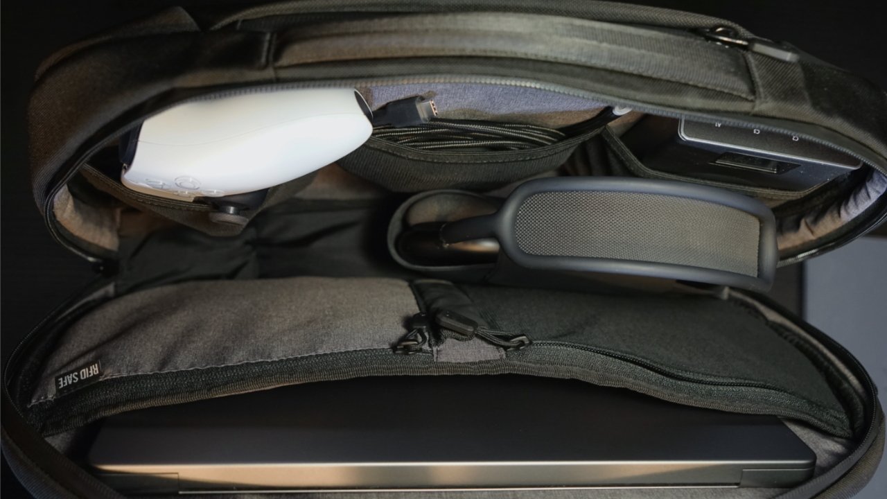 The interior of the Nomatic backpack showing a game controller, cables, a battery pack, AirPods Max, and an iPad Pro.