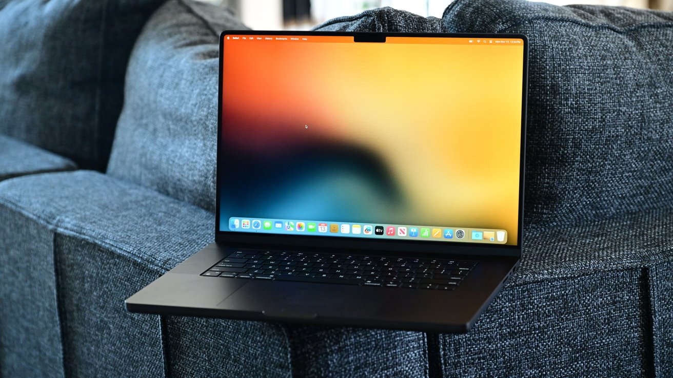 A 16-inch MacBook Pro sits on a blue-gray couch. It has a vibrant orange gradient wallpaper.
