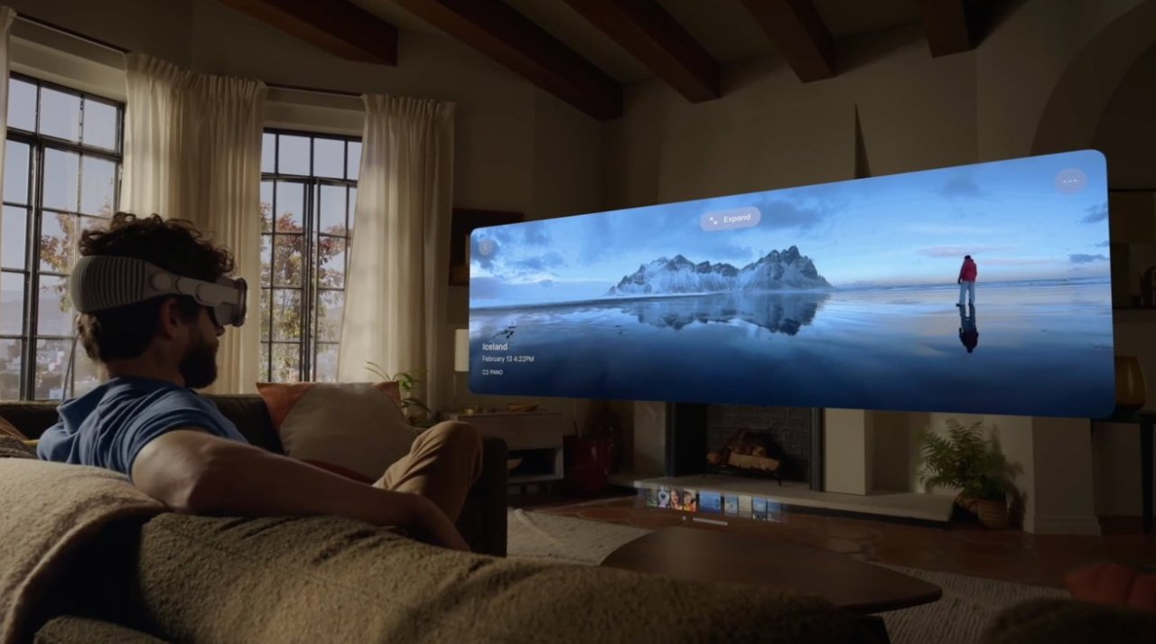 Man with VR headset sitting on a sofa, looking at a large panoramic screen showing a serene landscape with a reflective water surface.