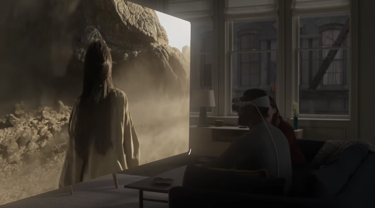 A person in a VR headset sits on a sofa while another figure stands facing a rocky landscape projected on a large screen in a sunny room.