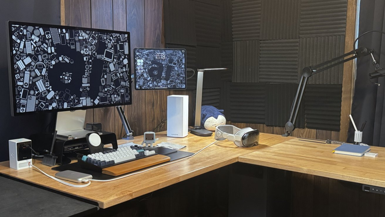 A desk with various computers and gadgets. Apple Vision Pro is visible next to a Studio Display, iPad Pro, keyboard, and other desk accessories.