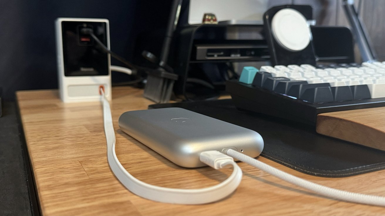 Apple Vision Pro battery lies on a desktop connected to a white USB-C cable. It extends from a black charging block and sits next to a keyboard, iPhone charger, and monitor stand.