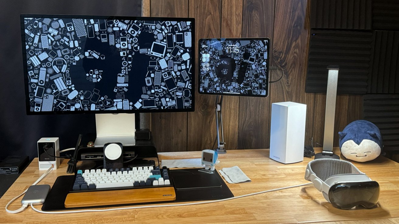 A desktop with a Studio Display, iPad Pro, keyboard, and other accessories. Apple Vision Pro is on the desk at the side.