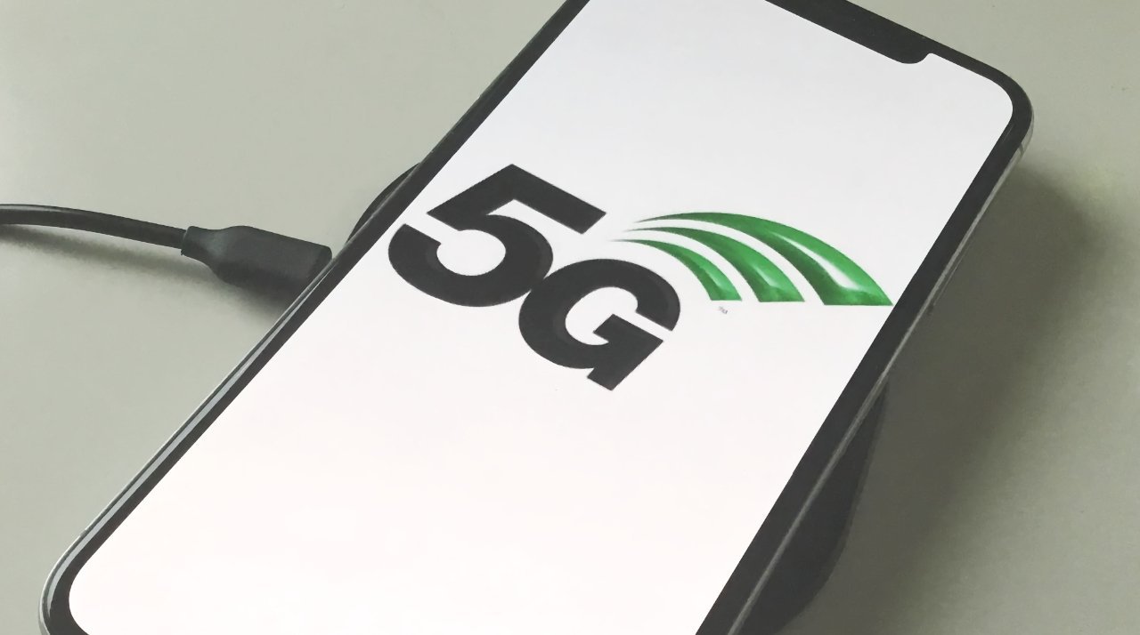 Smartphone with 5G logo on screen connected to a charging cable against a light background.