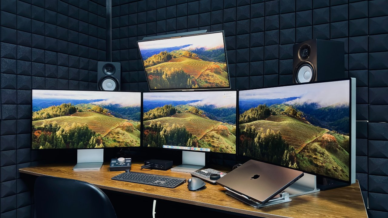 Modern workspace with three monitors displaying a scenic landscape, speakers, laptop, and peripherals on a desk with dark acoustic foam walls.