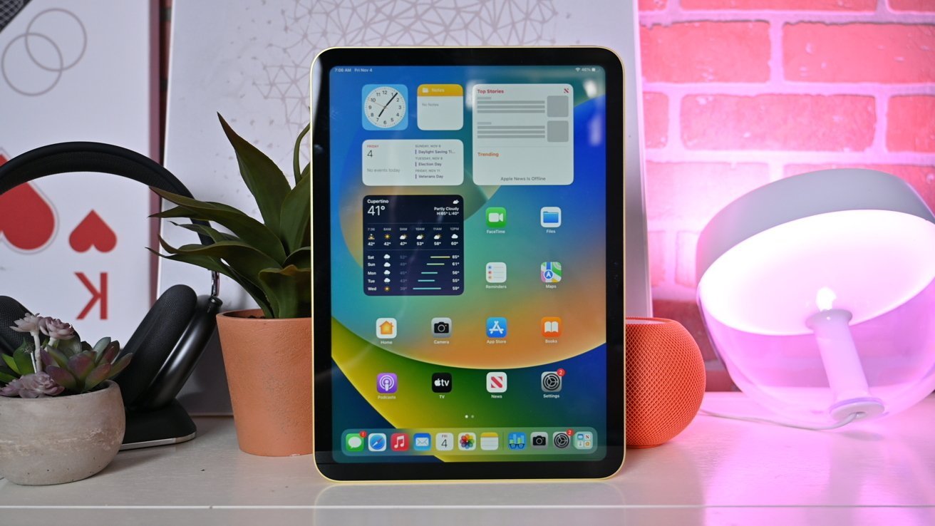 A yellow iPad 10th generation with widgets and apps displayed on a white desk with plants, speakers and a lamp against a red brick background.
