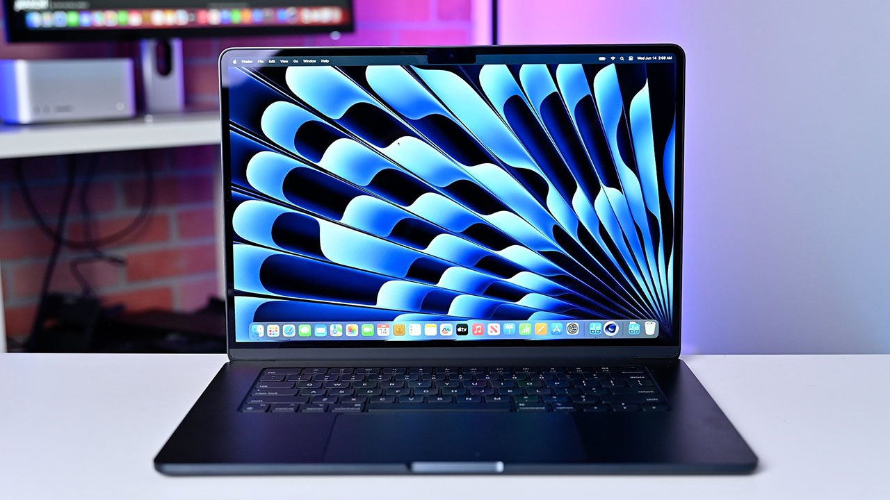 Midnight MacBook Air 15-inch laptop on a desk displaying a blue abstract wallpaper, with an out-of-focus colorful background.